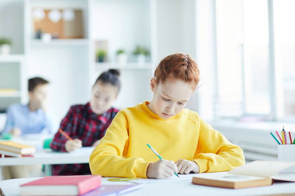 Roztomilá školka ve žlutém tričku, kde sedí na stole a dělá poznámky v učebnici v hodině - Fotografie, Obrázek
