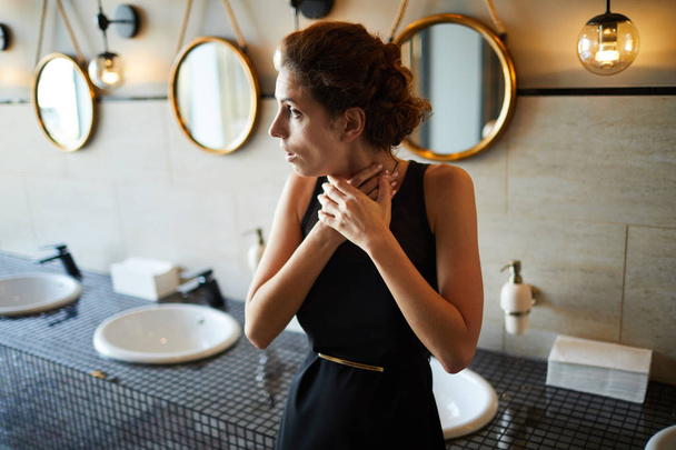 Jeune femme brune en robe noire élégante toucher sa gorge tout en se tenant dans les toilettes
 - Photo, image