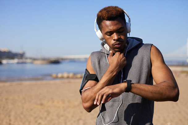 Hombre negro joven pensativo serio en auriculares con cable se conectan a un teléfono inteligente en el brazalete tocando el cuello mientras comprueba el pulso después de trotar
 - Foto, imagen