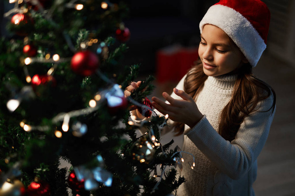 Маленькая девочка в шапочке Санты трогает украшения на ветке рождественской елки
 - Фото, изображение