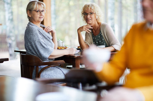 Dos curiosas hembras maduras en ropa casual sentadas en la cafetería y mirando al hombre bebiendo té junto a la mesa vecina
 - Foto, imagen