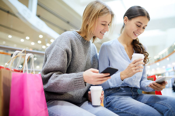 Cute shoppers with drinks and smartphones scrolling through notifications during break after shopping - Фото, изображение