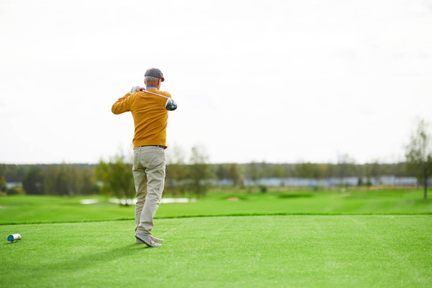 Back view of senior man in cap, yellow pullover and beige pants hit ball with golf club on green field - Photo, Image