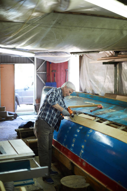 Senior master of shipbuilding drilling one of boards on unfinished vessel in workshop - Photo, Image