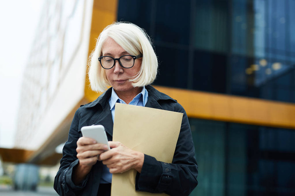 Loira mulher de negócios madura com documentos mensagens no smartphone ao ar livre
 - Foto, Imagem