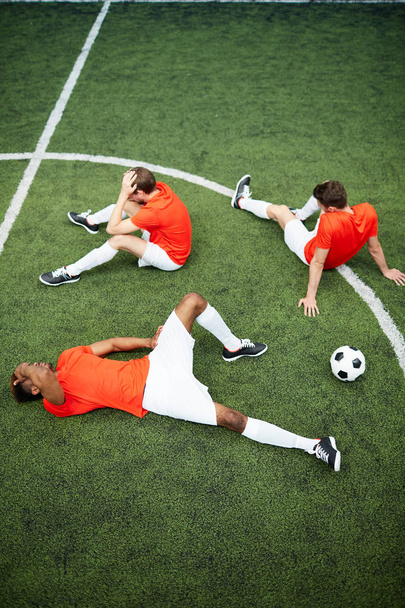 Three unlucky football players sitting and lying on green field after hard match or game - Foto, imagen