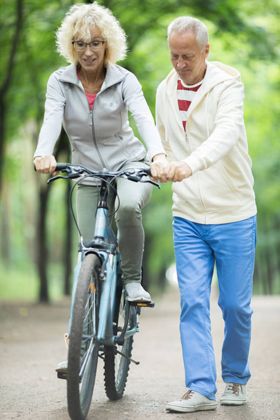 Hombre de edad que apoya a su esposa mientras le enseña a montar en bicicleta en el ocio
 - Foto, Imagen
