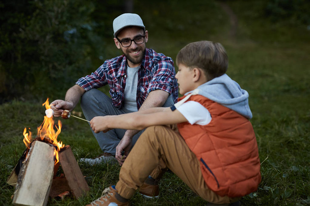 Sorrindo pai sentado perto do fogo junto com seu filho e torrando marshmallows
 - Foto, Imagem