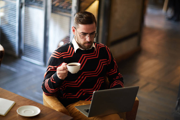 Joven con taza de café concentrándose en la red mientras está sentado en el sillón en la cafetería
 - Foto, imagen