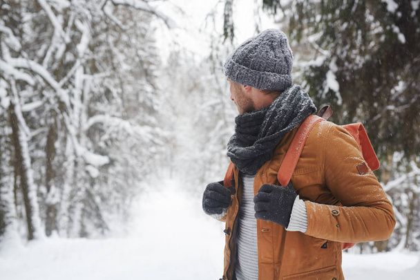 Pensive fiatal férfi hátizsákos a kötött kalap és sálat gazdaság kezeli a hátizsák, és nézett vissza az erdőben, miközben fontolgatják a téli természet - Fotó, kép