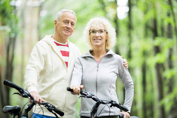 Στοργικό και αθλητικό ζευγάρι ηλικιωμένων που σας κοιτάζουν ενώ δροσεύουν στο πάρκο ή το δάσος με ποδήλατα - Φωτογραφία, εικόνα