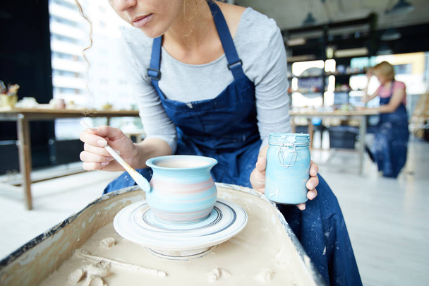 Female potter with jar of blue color sitting by rotating clay jug and painting it - Photo, Image