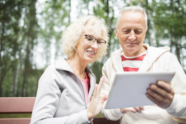 Счастливые современные пенсионеры в активах сидят в парке и разговаривают через видеочат в планшете
 - Фото, изображение