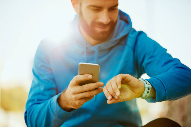Крупним планом молодий бородатий чоловік дивиться на розумний годинник і тримає смартфон в іншій руці
. - Фото, зображення