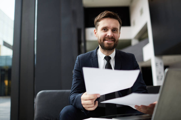 Щасливий бородатий бізнесмен з паперами, які дивляться на вас під час роботи в сучасному офісі
 - Фото, зображення