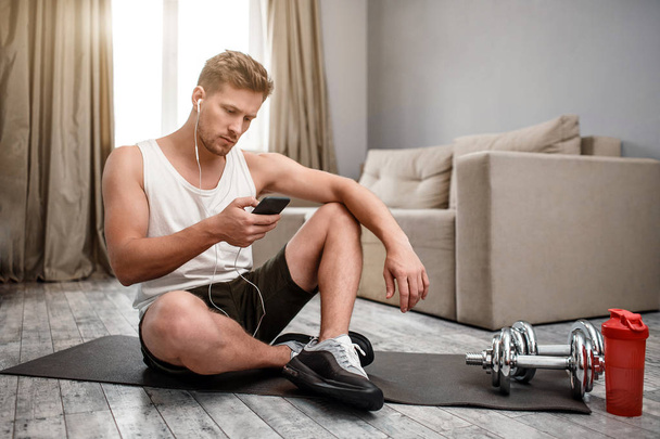 Young well-built man go in for sports in apartment. He sit on carimate and hold phone. Guy listen to music through earphones. Dumbbells with red water bottle on right. - Fotó, kép