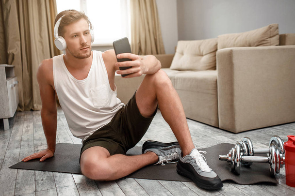 Young well-built man go in for sports in apartment. He sit on carimate floor and take selfie. Guy wear headphones. Relax. Take pause. - Fotó, kép