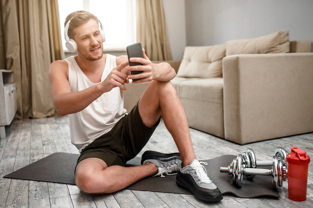 Young well-built man go in for sports in apartment. Happy positive guy sit on carimate on floor and take selfie. Dumbbells and water bottle beside. - Photo, image