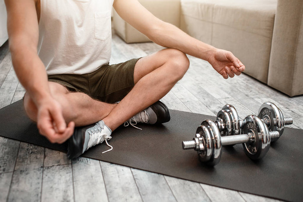Young well-built man go in for sports in apartment. He sit on carimate with legs crossed and meditate. Dumbbells lying in front of him. Cut view. - Фото, изображение