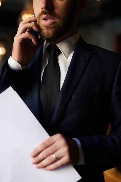 Κοντινό-up του σίγουρος μουσάτο επιχειρηματία σε μοδάτο κοστούμι κρατώντας έγγραφα και να κάνει επιχειρήματα, ενώ μιλάει στο κινητό τηλέφωνο - Φωτογραφία, εικόνα