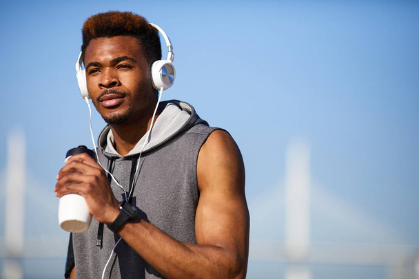 Serious pensive sporty African guy with mustache listening to music via modern headphones and drinking protein beverage while refreshing after outdoor training - Foto, Imagen