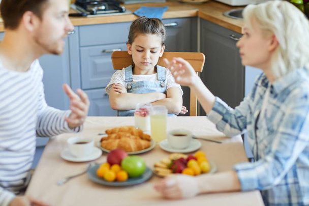 Aufgebrachtes kleines Mädchen sitzt am gedeckten Tisch, während sich ihre Eltern beim Frühstück streiten - Foto, Bild