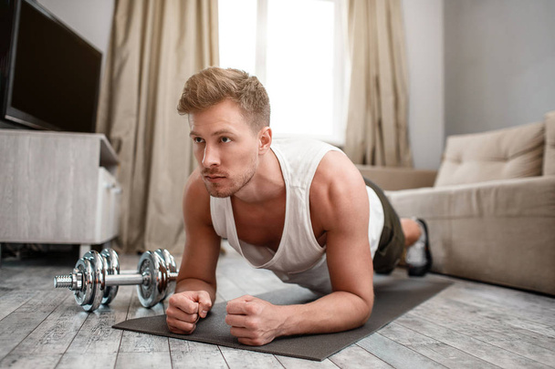 Young well-built man go in for sports in apartment. He stand in plank position on carimate and look forward. Strong and muscular. Dumbbells lying beside. - Fotoğraf, Görsel