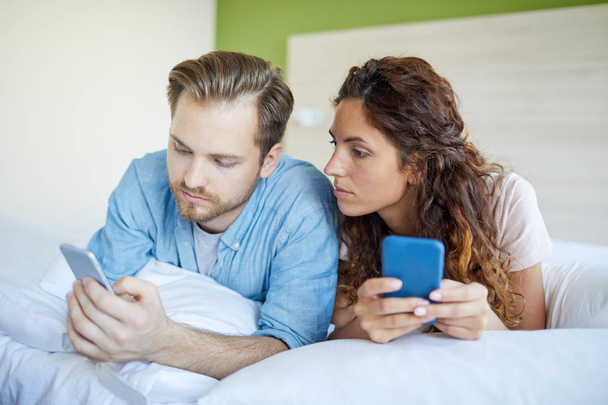 Νεαρό ζευγάρι ξαπλωμένο στο κρεβάτι και ψάχνουν ή στέλνουν μηνύματα στα smartphones τους - Φωτογραφία, εικόνα
