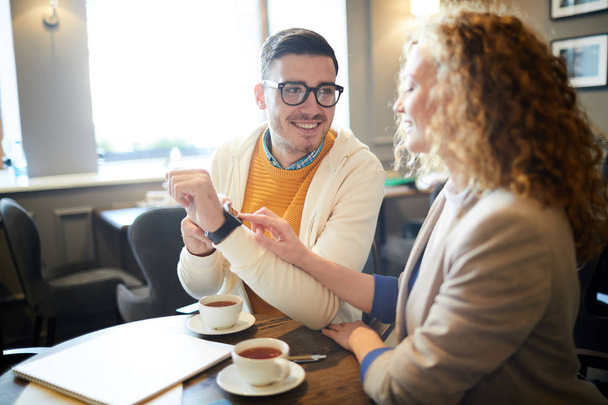 Mladý ležérní muž ukazuje svůj nový smartwatch na svého kolegu nebo přítelkyni šálek čaje v kavárně - Fotografie, Obrázek