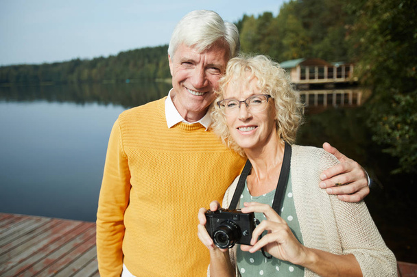 glückliche Seniorentouristen in modischer Kleidung, die sich umarmen und in die Kamera lächeln, während sie auf der Seebrücke stehen und in die Kamera schauen, ruhen sie auf dem Land - Foto, Bild