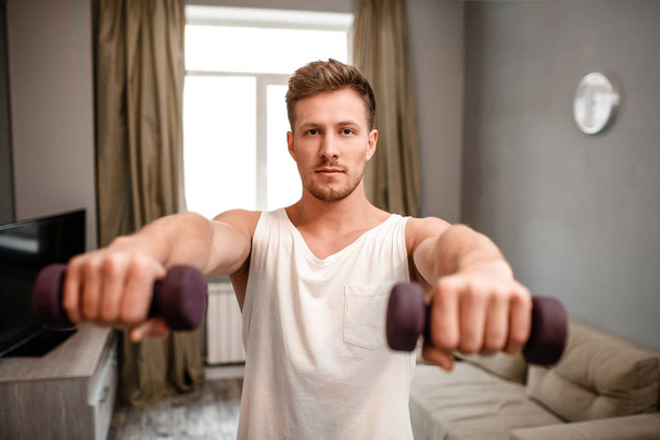 Young well-built man go in for sports in apartment. He hold dumbbells in front of himself and look on camera. Strong and muscular young man has workout. - Zdjęcie, obraz