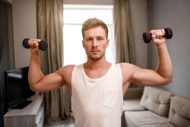 Young well-built man go in for sports in apartment. He does shoulder press using dumbbells. Guy look on camera serious. Strong and muscled guy. - Foto, Imagem