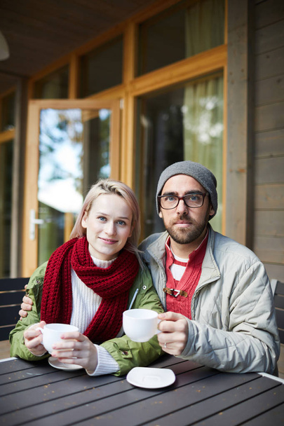 Portrait of young attractive Caucasian couple sitting at wooden table on terrace, holding cups of hot tea and looking at camera - Foto, imagen