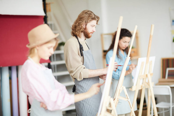 Jovem de avental em pé à beira do cavalete entre colegas de grupo e pintura na lição no estúdio de arte
 - Foto, Imagem
