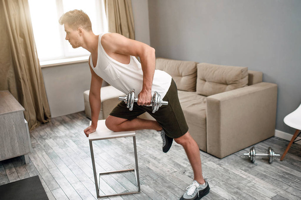 Young well-built man go in for sports in apartment. He does dumbbell row exercise. Concentrated strong muscled young man. - Фото, изображение