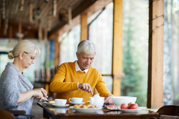 Zawartość Starsza para w casual Odzież siedzi przy stole i jedzenia żywności, podczas gdy śniadanie na ganek wsi domu - Zdjęcie, obraz