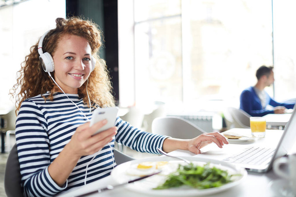 Šťastná krásná holčička s kudrnatými vlasy, které sedí u stolu, a vybírá si na smartphone, když pracuje s laptopem a snímá v moderní kavárně - Fotografie, Obrázek