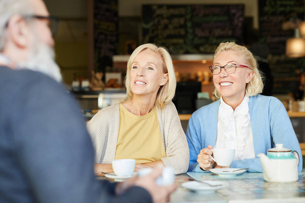 zwei hübsch gealterte, fröhliche Frauen in klugem, lässigem Blick auf einen älteren Mann während ihres Gesprächs im Café - Foto, Bild