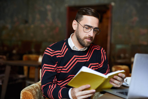 Молодий серйозний чоловік в окулярах читає книгу, проводячи дозвілля в кафе
 - Фото, зображення