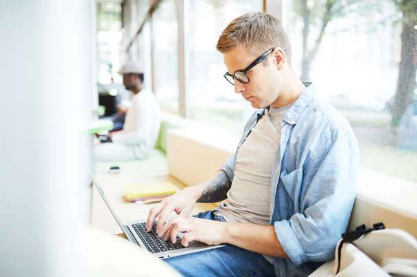 Випадковий молодий чоловік друкує на клавіатурі ноутбука, сидячи біля вікна в коледжі
 - Фото, зображення