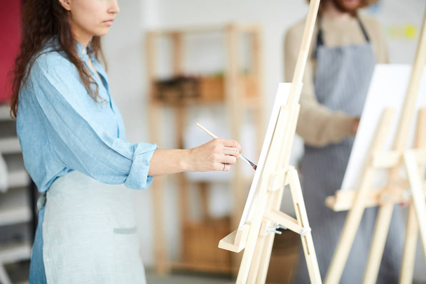 Stüdyodaki modern sanatların sehpasında boya fırçası ile resim yapan genç bir kadın. - Fotoğraf, Görsel