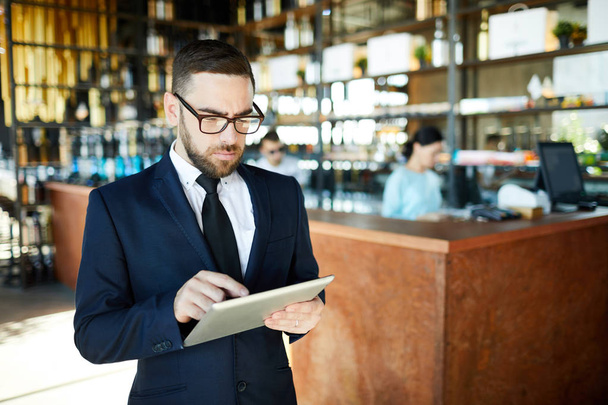 Liikemies, jolla on tabletti, keskittyy etsimään tietoja verkosta työskennellessään kahvilassaan
 - Valokuva, kuva