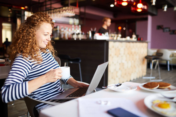Pozitív vonzó göndör hajú, fiatal női szabadúszó marketingszakértő iszik kávét és ellenőrzési információk laptop munka közben a kávézóban - Fotó, kép