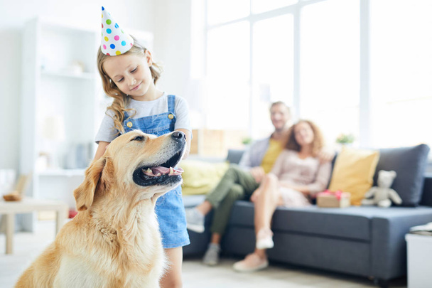 Lindo niño en la gorra de cumpleaños de pie junto a su perro y abrazarlo con los padres relajantes en el fondo
 - Foto, Imagen