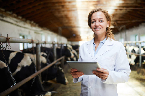 Portrait taille haute de gaie vétérinaire femme souriant regardant la caméra tout en utilisant une tablette numérique debout dans étable de ferme laitière moderne, espace de copie
 - Photo, image