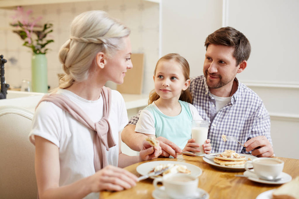 Conteúdo bela família sentada à mesa de jantar e conversando enquanto come deliciosas panquecas caseiras na cozinha doméstica
 - Foto, Imagem