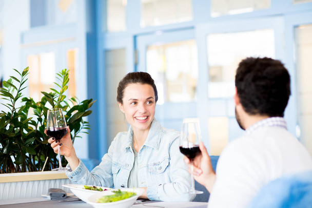 Щаслива молода жінка з келихом червоного вина дивиться на свого чоловіка і розмовляє з ним під час обіду в ресторані
 - Фото, зображення