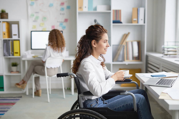 Trabajador de oficina joven en silla de ruedas tomando té o café por el lugar de trabajo mientras ve webcast
 - Foto, Imagen