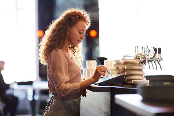 Serious pensive attractive curly-haired waitress standing at bar counter and using pencil while adding order in restaurant POS - Foto, imagen