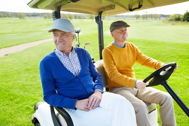 Dos amigos mayores sentados en un coche de golf mientras uno de ellos conduce para jugar al golf
 - Foto, Imagen